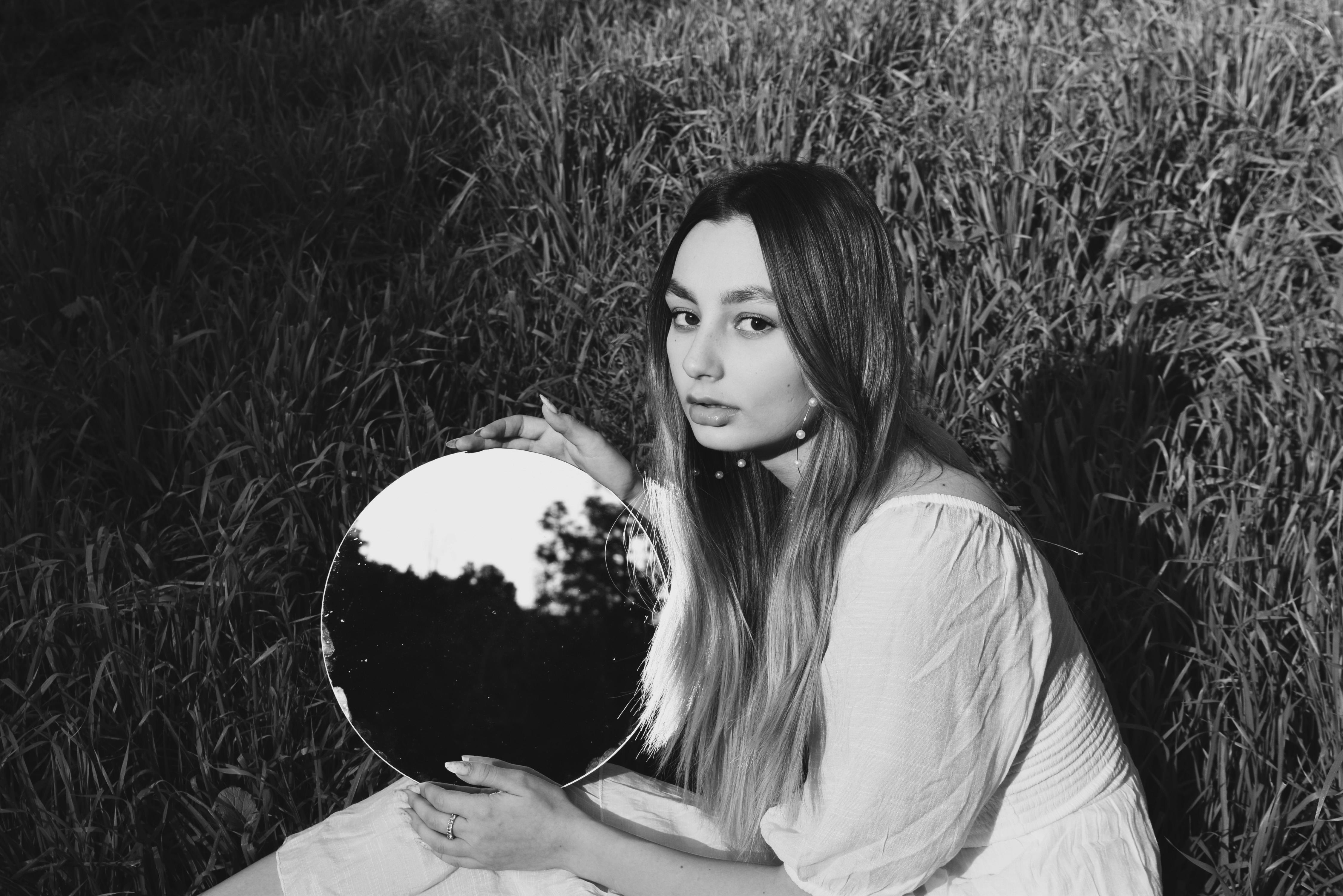 Artistic black and white portrait of a woman with long hair holding a mirror outdoors.